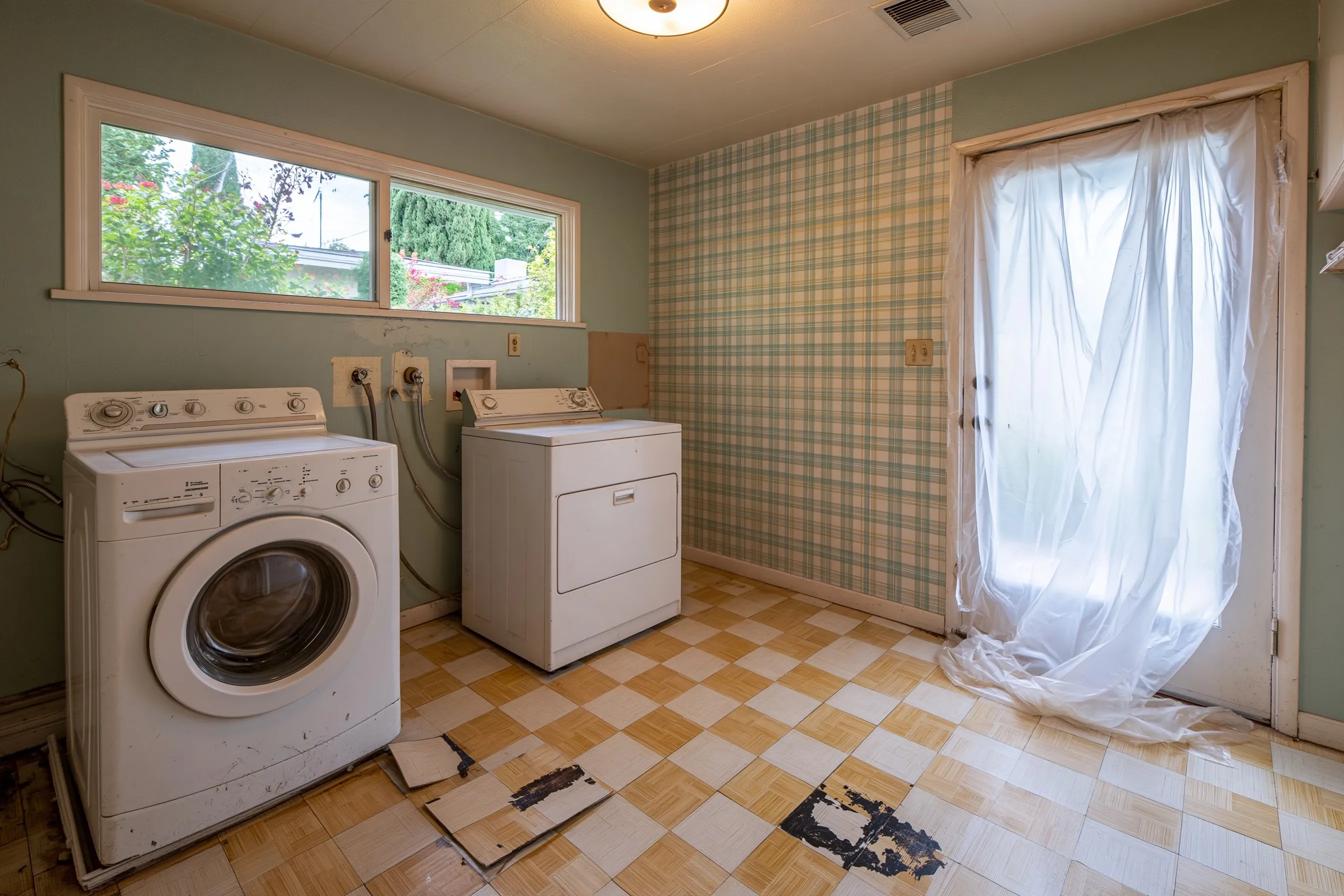 A 1960s laundry room emptied of its washer and dryer, with 9-inch floor tiles lifted to show black adhesive