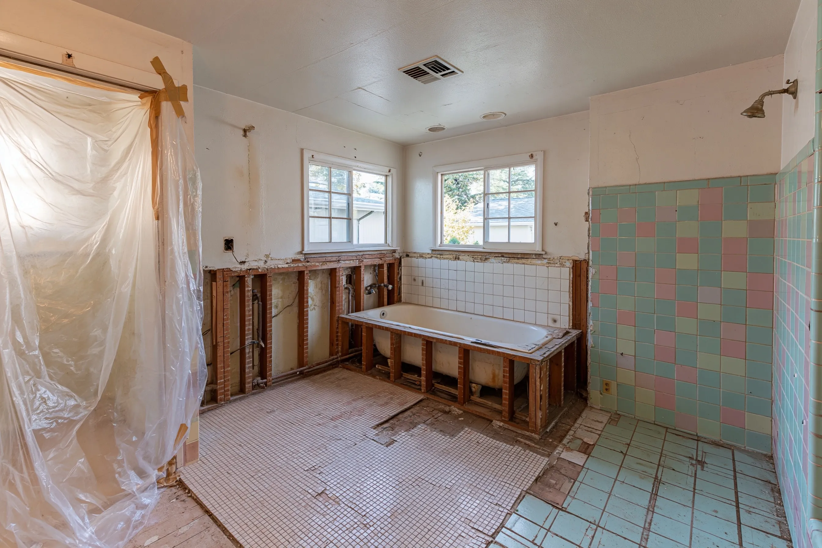 A 1950s bathroom opened for a remodel, with the tub wall cut out, floor tile lifted and plastic hung